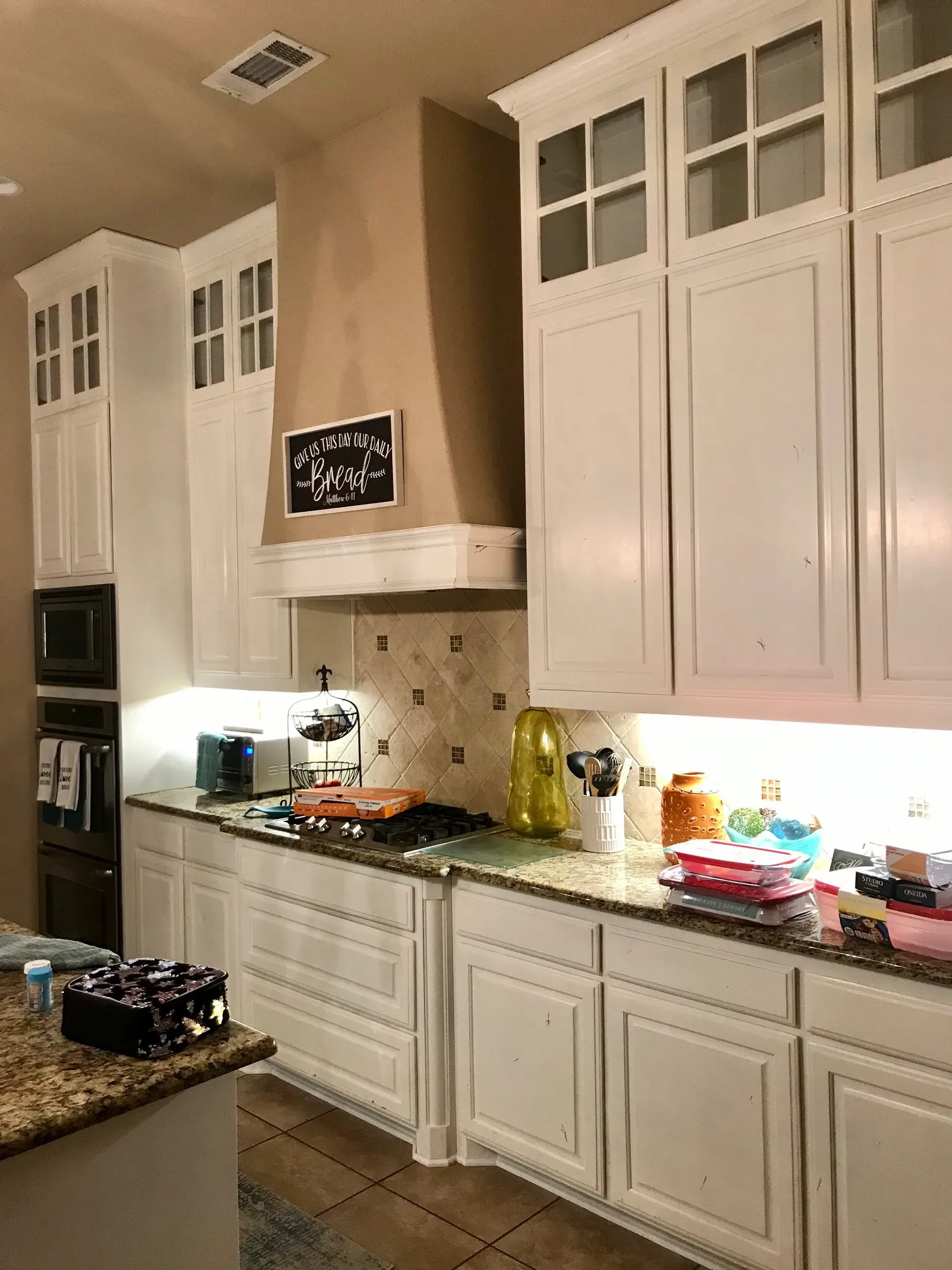 kitchen cabinets with glass panels and granite countertops, featuring an arched vent hood with a 'Give us this day our daily bread' sign