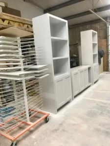 White custom cabinetry, including shelving units and lower cabinets, being dried in a workshop